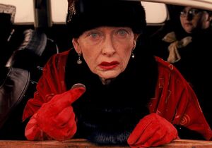 Old Tilda Swinton in red as Madame D. with coin